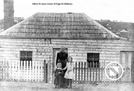 A woman and two children stand in front of a small wooden house with a picket fence.